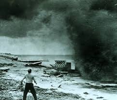 Historical photograph of a person near waterfront structures with a large plume of water and spray behind them.